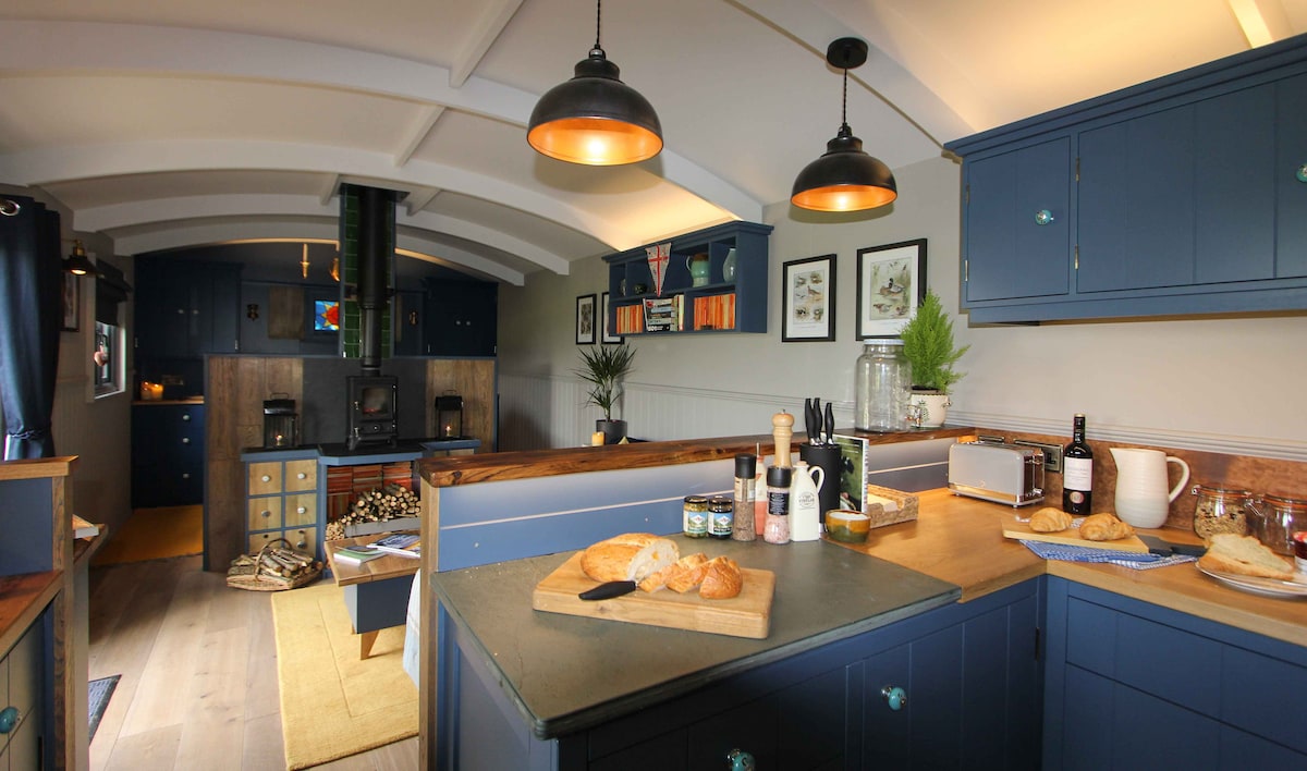 A welcoming kitchen area features blue cabinetry and a spacious countertop, adorned with freshly baked items. Pendant lights provide soft illumination, while the wood stove and various decor elements complete the cozy atmosphere. A functional layout is evident, ensuring a comfortable cooking experience.
