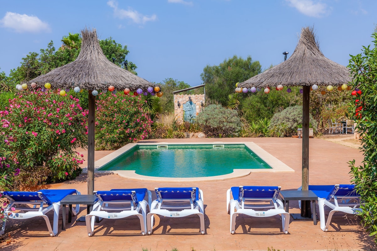 A serene outdoor space features a private swimming pool surrounded by lush greenery. Two shaded areas provide lounge seating under thatched roofs, with colorful lights strung above. Sun loungers are positioned near the pool, offering a view of the tranquil setting.