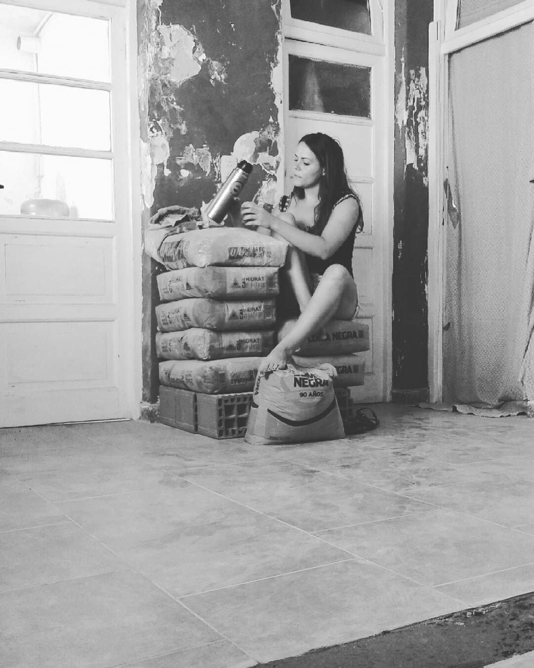 A seated individual is positioned on a stack of bags sitting on a tiled floor. The background shows a partially renovated room with exposed walls and natural light coming from a window. Construction materials are arranged nearby, indicating ongoing home improvements.