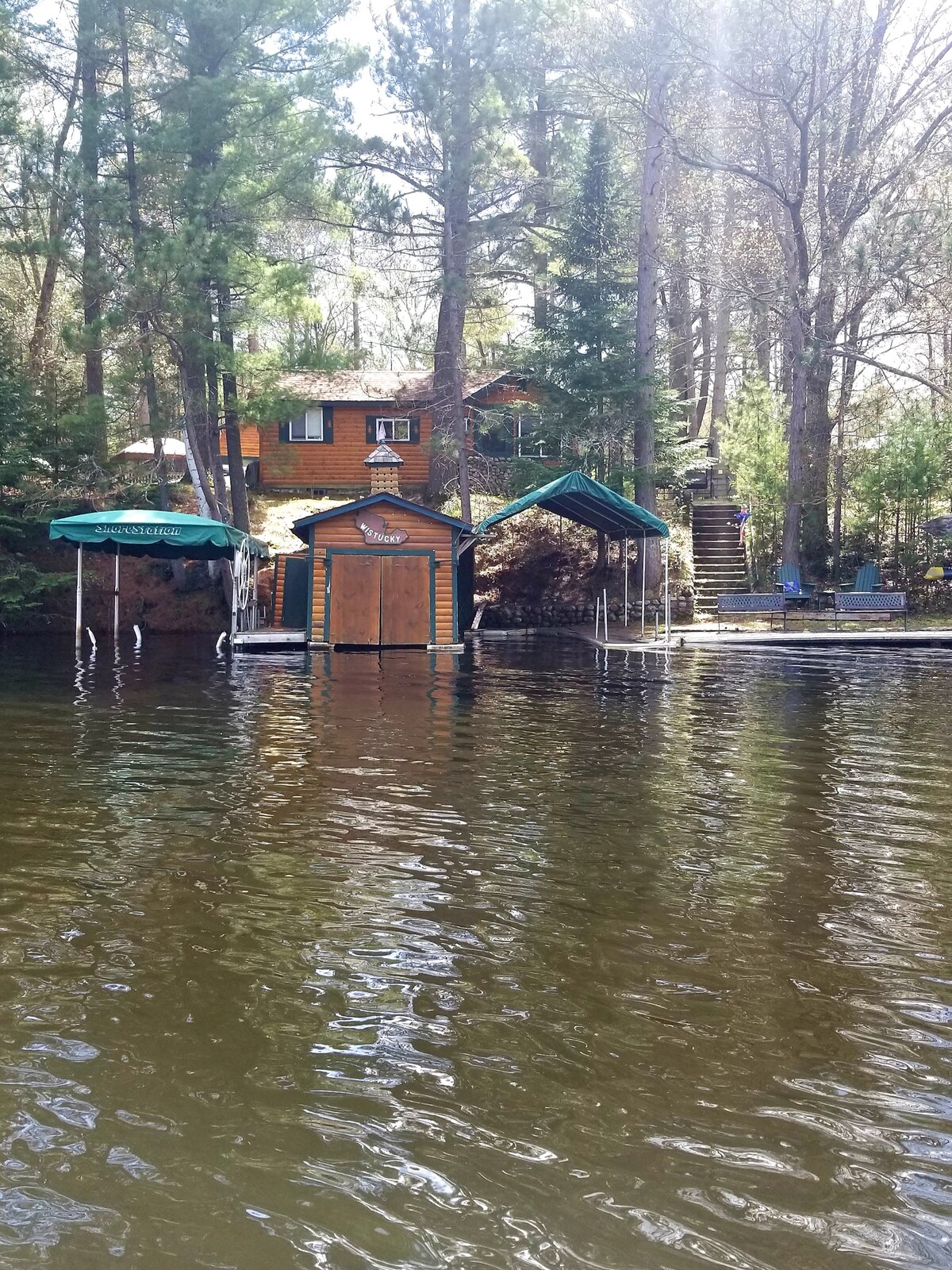 The image depicts a serene lakeside scene featuring a rustic cabin surrounded by tall trees. A covered boat slip is visible in the foreground, alongside a grassy area with chairs. Natural light filters through the trees, enhancing the tranquil atmosphere.