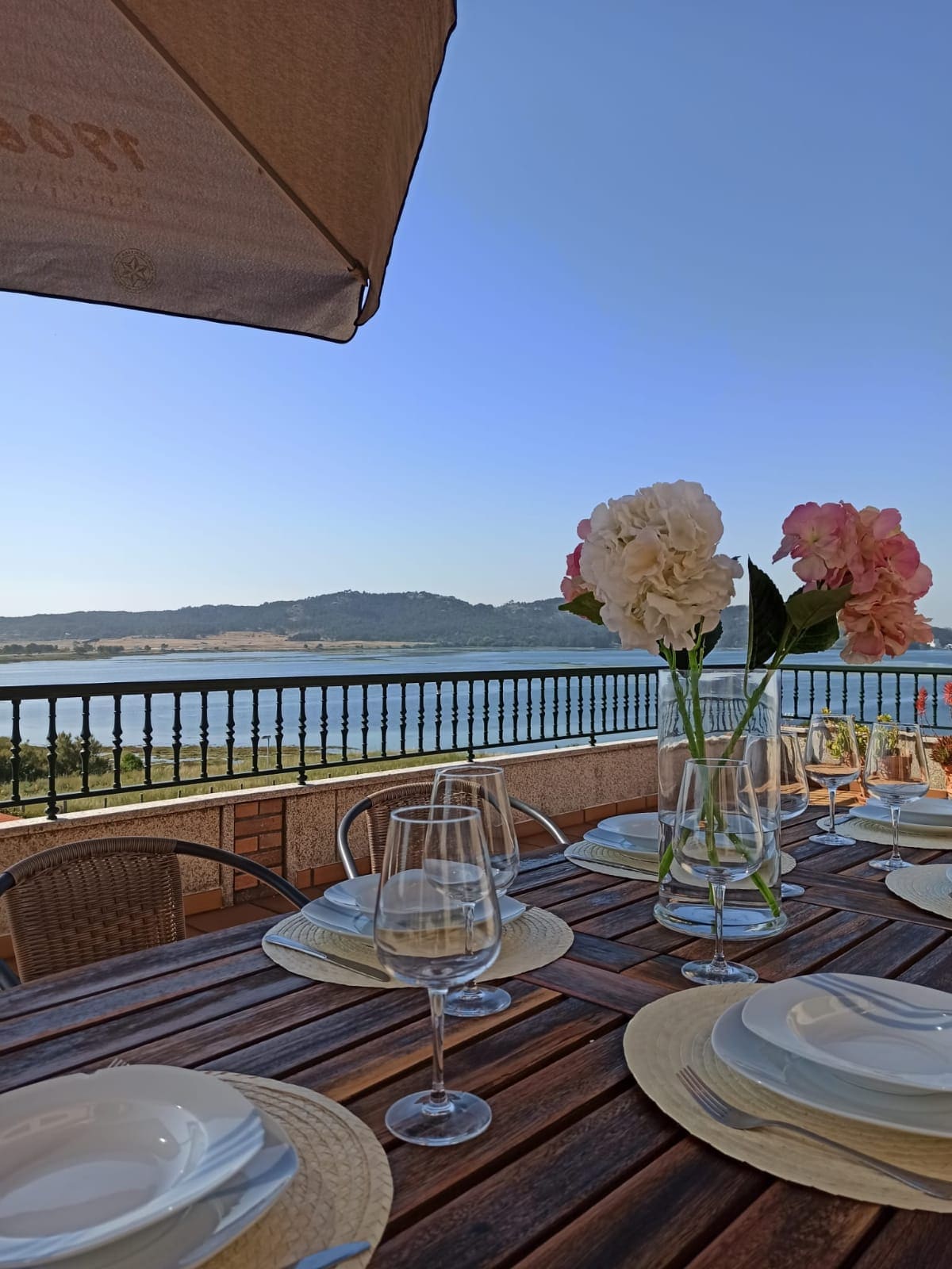 A dining table is set on a terrace, featuring multiple plates and glasses arranged for a meal. Floral centerpieces add a touch of nature. Scenic views of the water and distant hills provide a serene backdrop, complemented by a clear blue sky.
