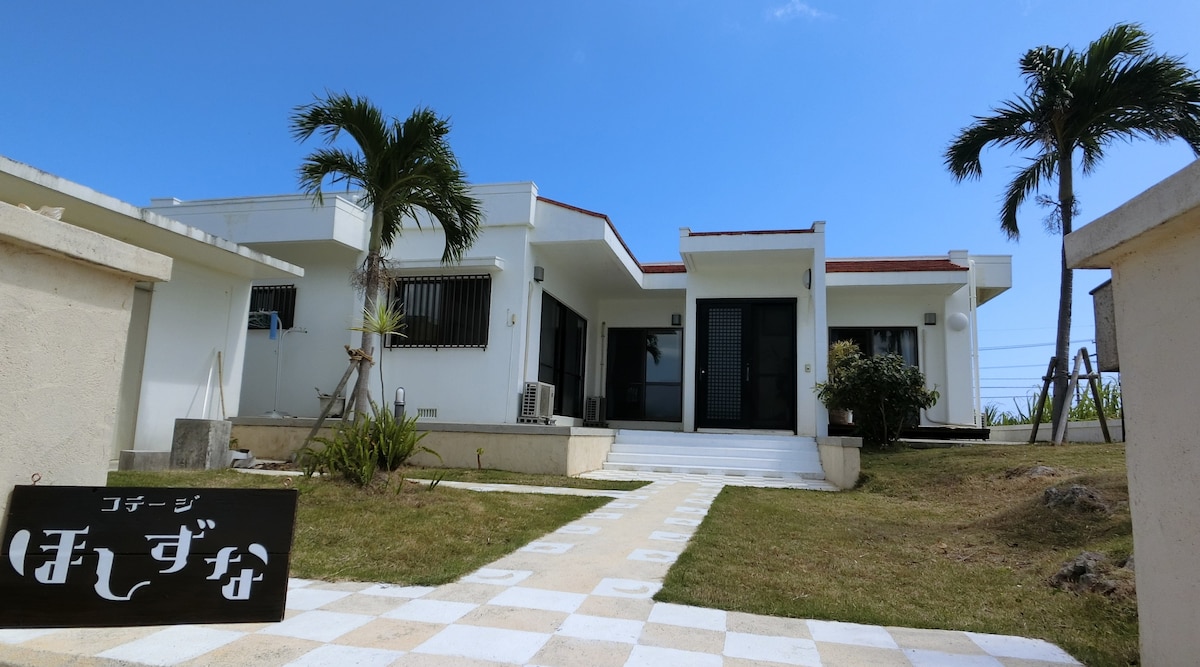 A modern, single-story building is shown with a clean white facade, surrounded by a grassy area. Pathways of light and dark tiles lead to the entrance, where large doors are framed by windows on each side. Tropical plants add greenery to the setting.