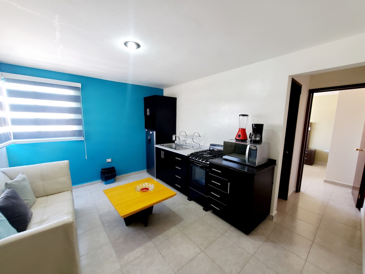 A compact living space features a smooth tiled floor and a bright blue accent wall. A light-colored sofa is positioned beside a yellow coffee table. The kitchenette includes a stove, refrigerator, and blender, while natural light enters through a window with modern striped shades.