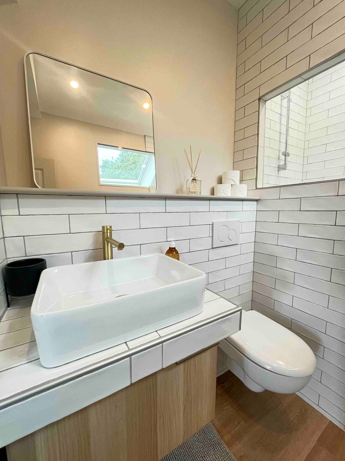 The modern bathroom features white tiled walls with a contrasting wooden vanity. A square sink sits atop the vanity, accompanied by a stylish faucet. Natural light enters through a small window, illuminating the space, while elegant decor items are arranged neatly on the countertop.
