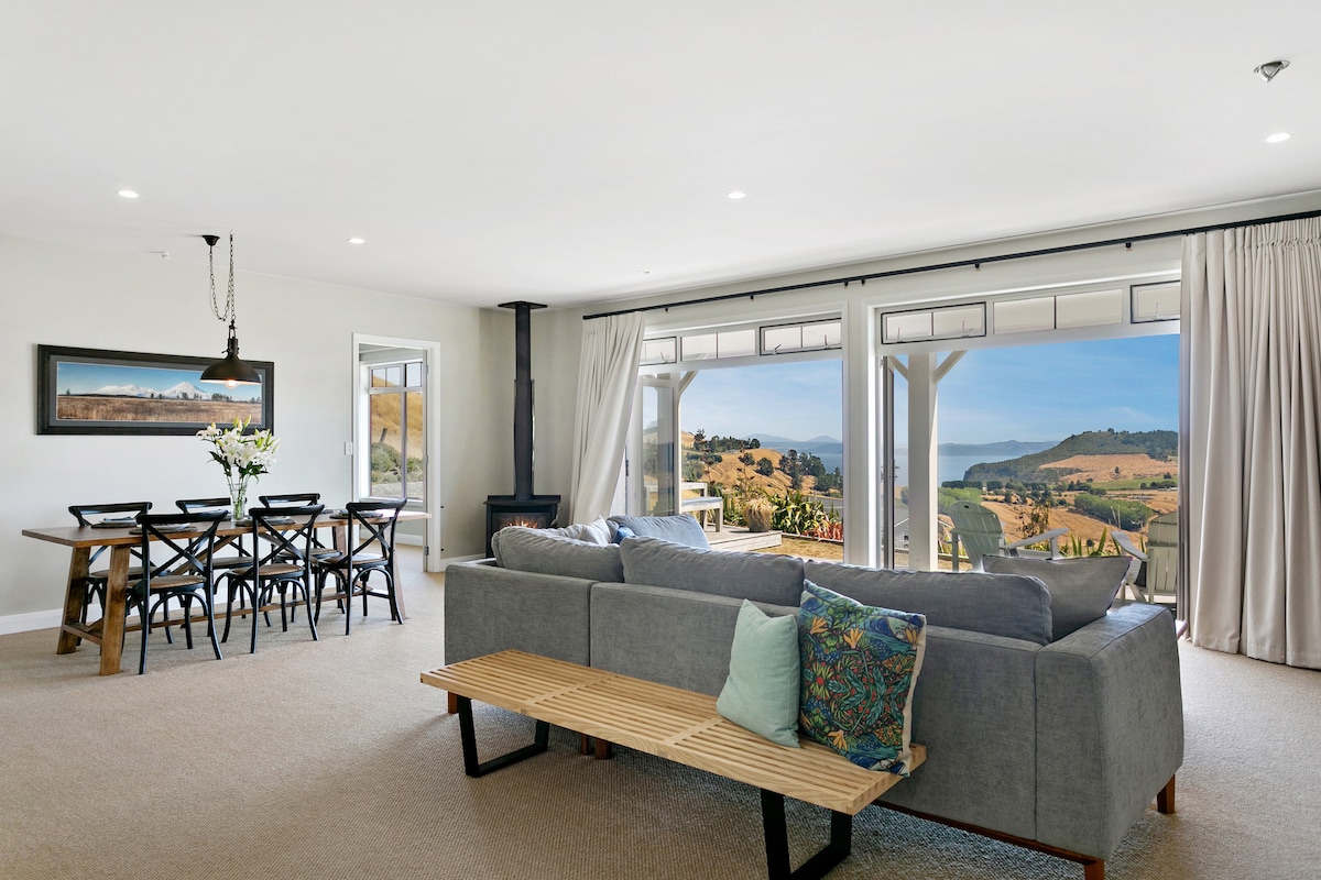 The open plan living and dining area features large glass sliding doors that frame expansive views of the countryside and lake. A cozy sofa with colorful cushions is positioned in front of a wooden coffee table. A dining table with black chairs is set for gatherings.