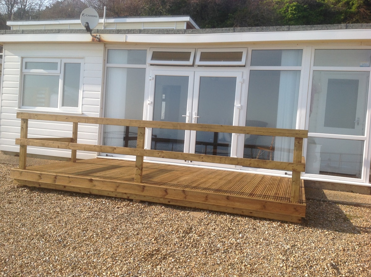 A wooden deck extends from the front of the villa, providing an accessible entry point. Large French doors are visible, leading into the interior. A gravel surface surrounds the deck, and the villa is positioned against a backdrop of greenery.