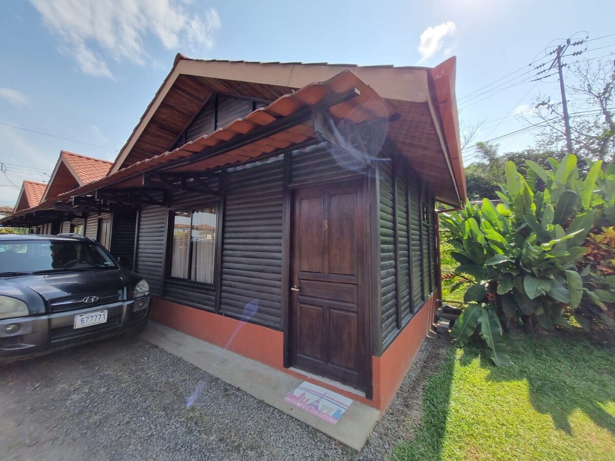A single-story cabin is shown with a wooden structure featuring dark wooden siding and a pitched roof. A welcoming entrance door is visible, framed by two windows that permit natural light. Lush greenery and a parked car are seen in the foreground.