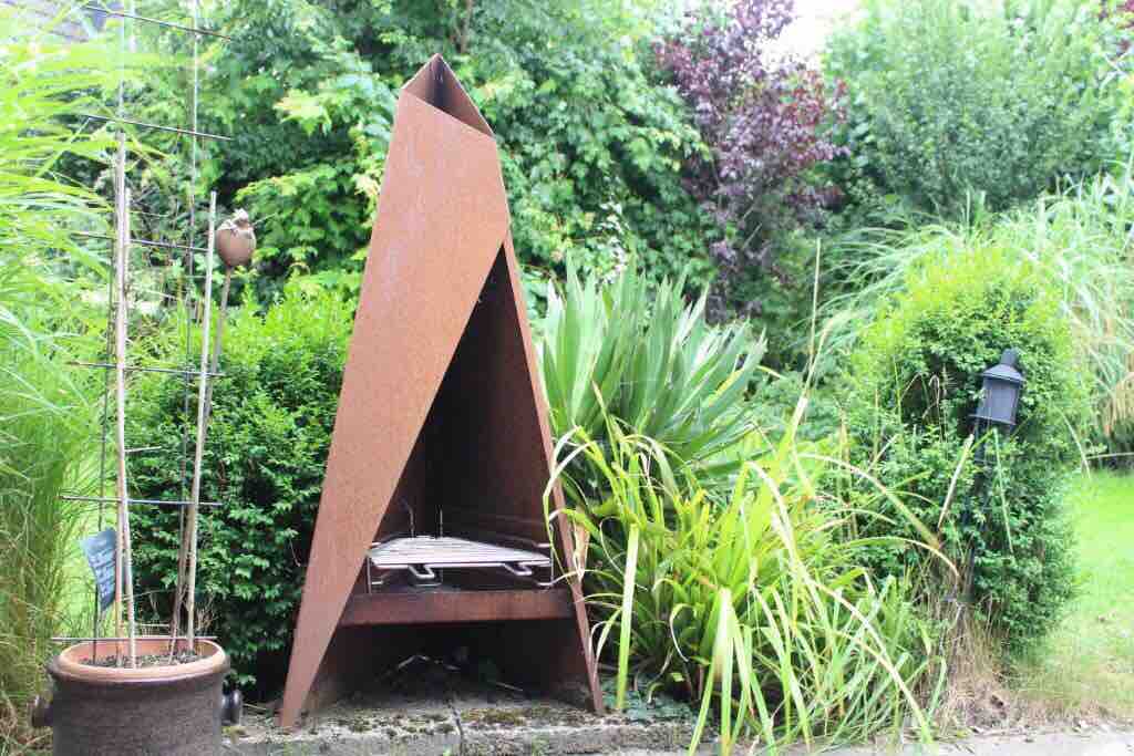A triangular garden grill with a rusted metal finish is positioned amidst lush greenery. Surrounding plants include tall grass and dense shrubs, creating a natural setting. The grill is designed to function as both a barbecue and a fire pit, enhancing the outdoor experience.