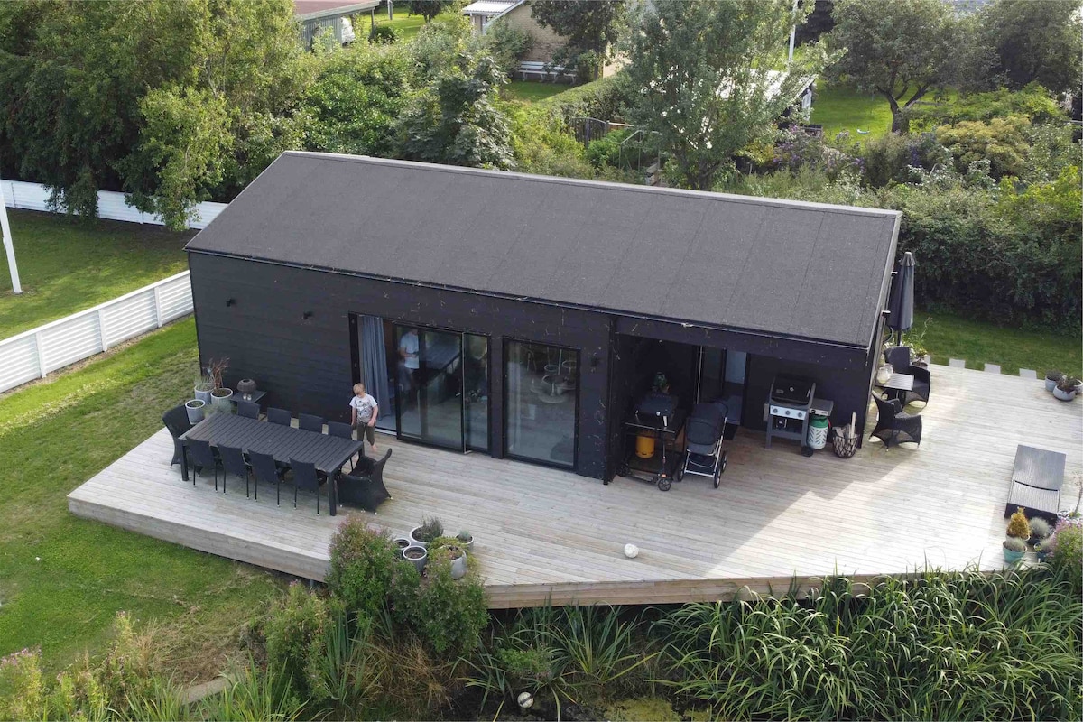 The vacation house features a sleek black exterior with expansive glass doors opening to a wooden deck. A large outdoor dining table is visible, surrounded by seating. Lush greenery and a tranquil water feature create a serene environment.