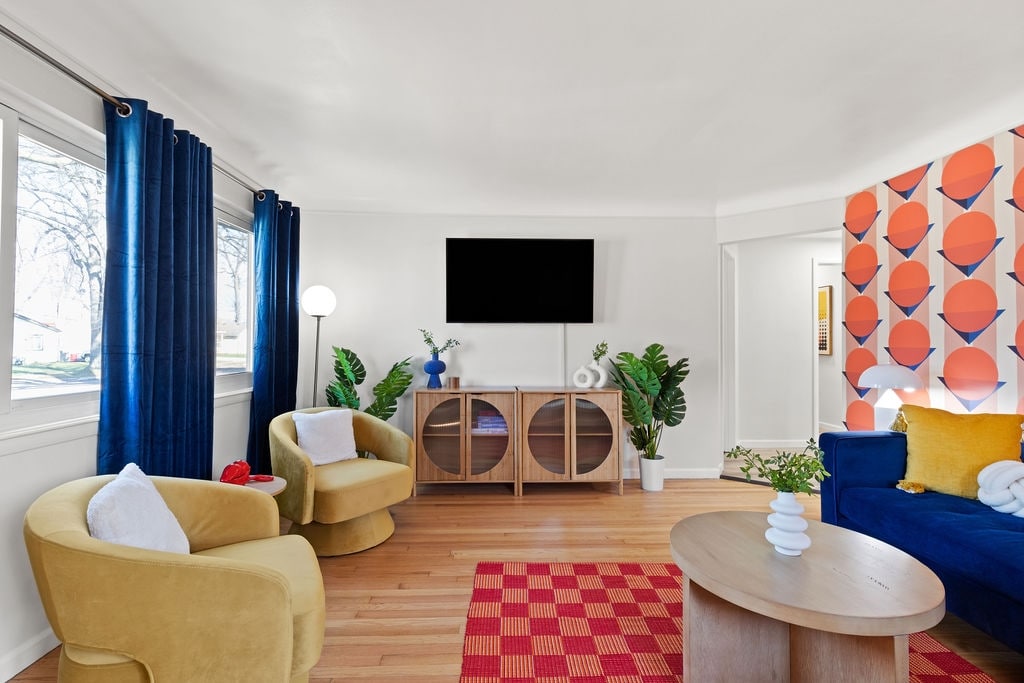 The living area features a vibrant blue sofa paired with two yellow accent chairs, creating a cozy seating arrangement. A round coffee table sits at the center, while a textured orange and red rug adds warmth. Natural light streams through large windows, complemented by blue curtains.