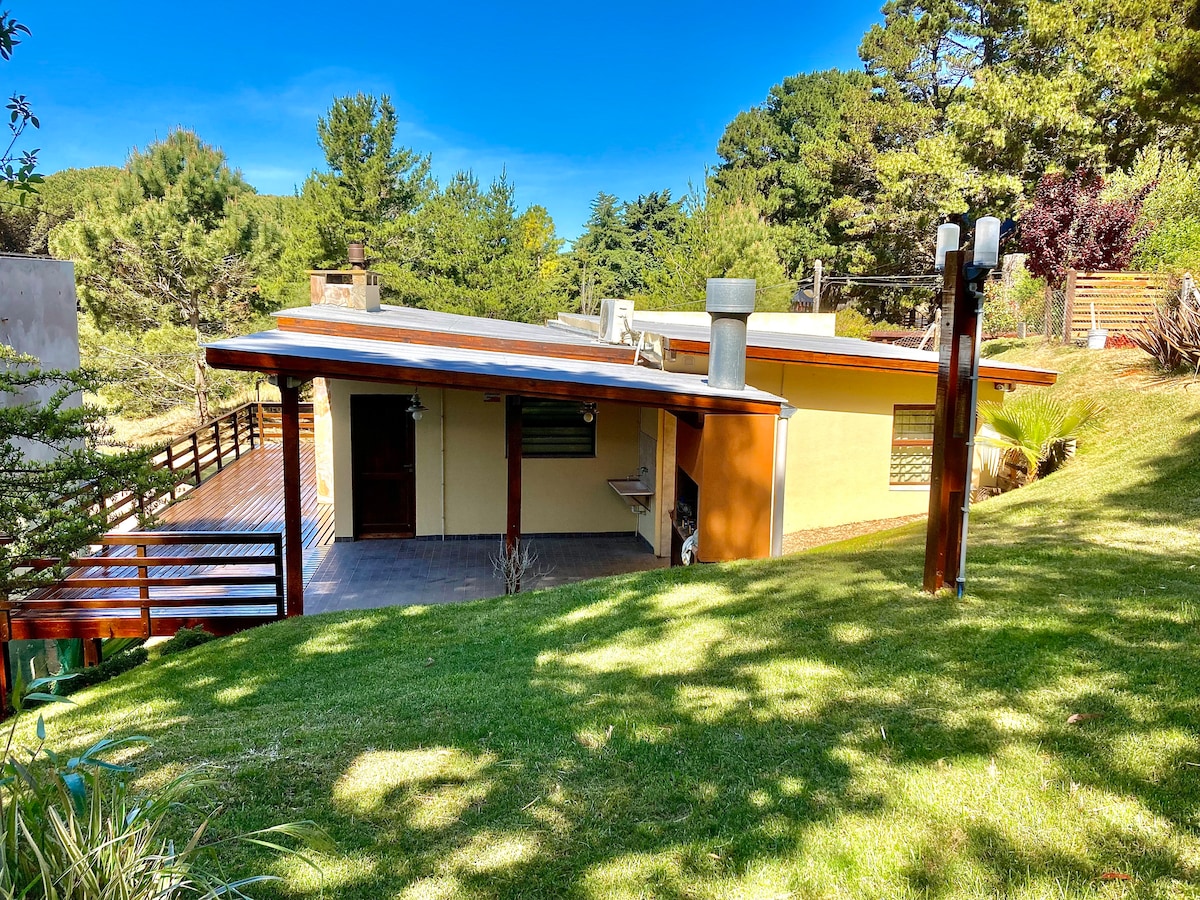 A single-story house nestled in a lush green landscape is shown. The structure features large windows and a covered porch with wooden beams, surrounded by trees. A manicured lawn extends in front, offering a peaceful outdoor space.