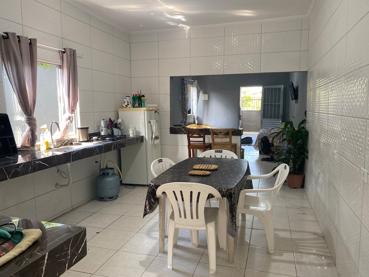 A bright kitchen area is equipped with a table surrounded by chairs, and a large window letting in natural light. A gas cylinder is placed near the counter, alongside a refrigerator. The walls have a clean, tiled finish, enhancing the spacious feel of the room.