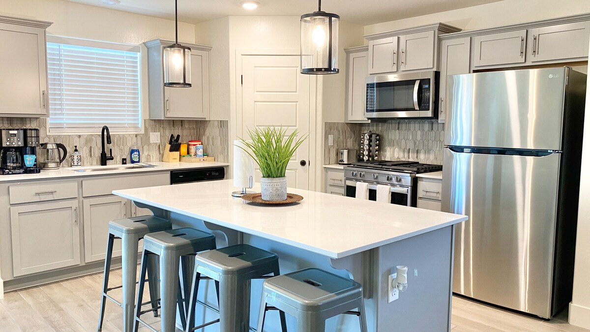 The kitchen showcases modern appliances, including a stainless steel refrigerator and a gas stove. Grey cabinetry complements the white countertops, with a central island featuring three metal stools. Natural light enters through a window, enhancing the airy feel of the space.
