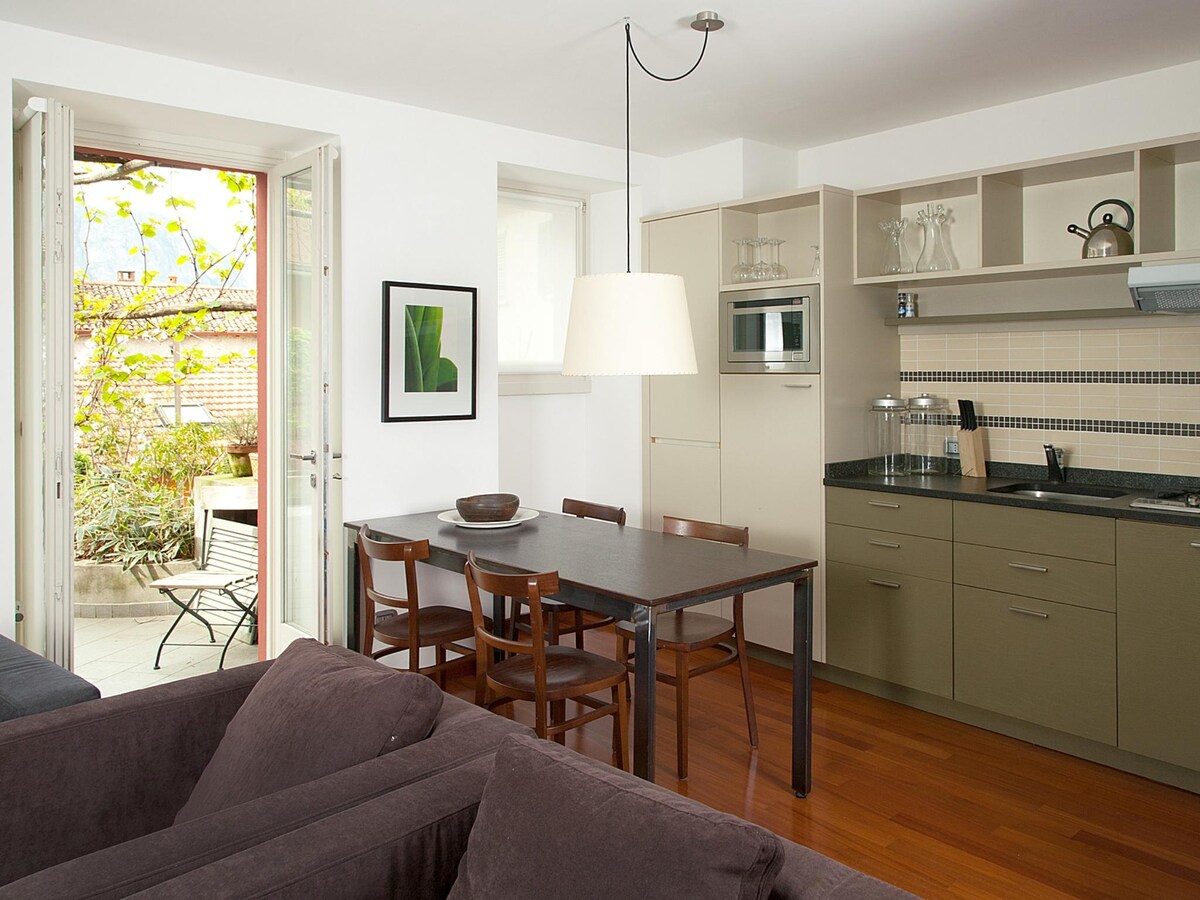 An open kitchen area is displayed, featuring modern cabinets and a dark countertop. A dining table with four wooden chairs is positioned centrally. The space is brightened by a large window, which leads to a private terrace with green plants visible outside.