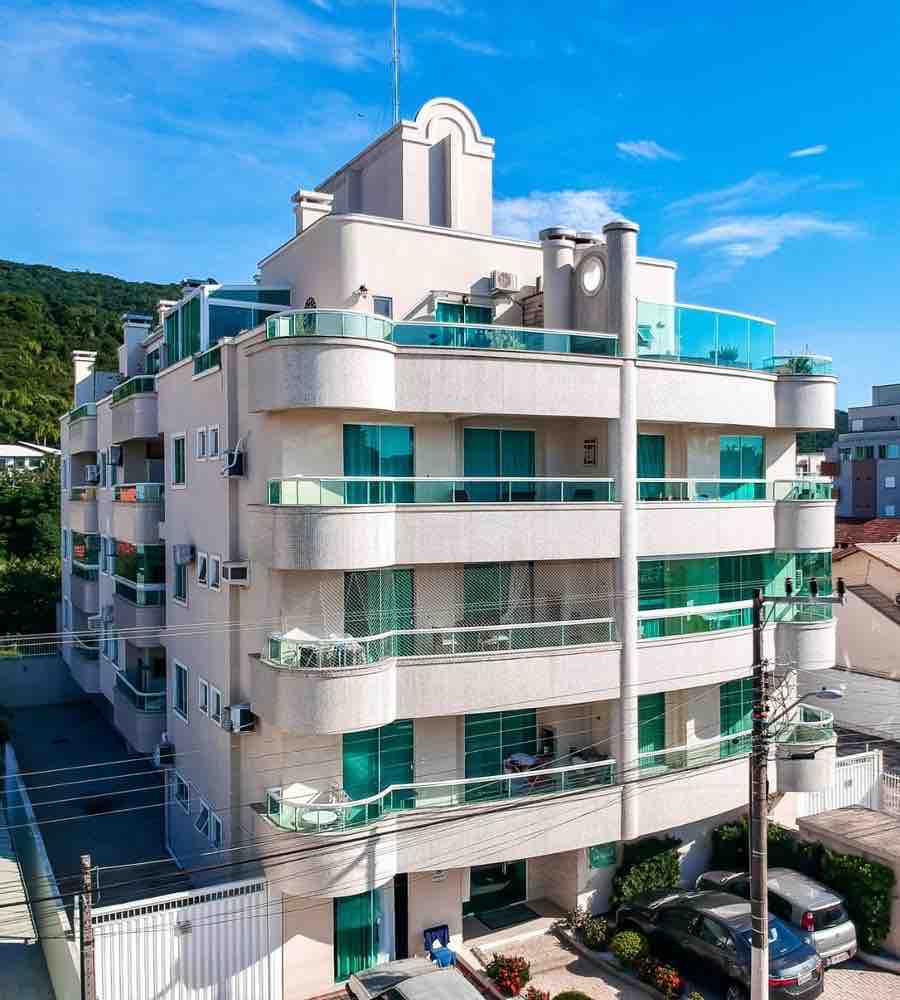 A contemporary four-story building is showcased with a clean design featuring multiple balconies adorned with glass railings. Air conditioning units are visible, and the exterior is complemented by lush greenery and a well-maintained garden area. The sky above is clear, enhancing the building's appeal.