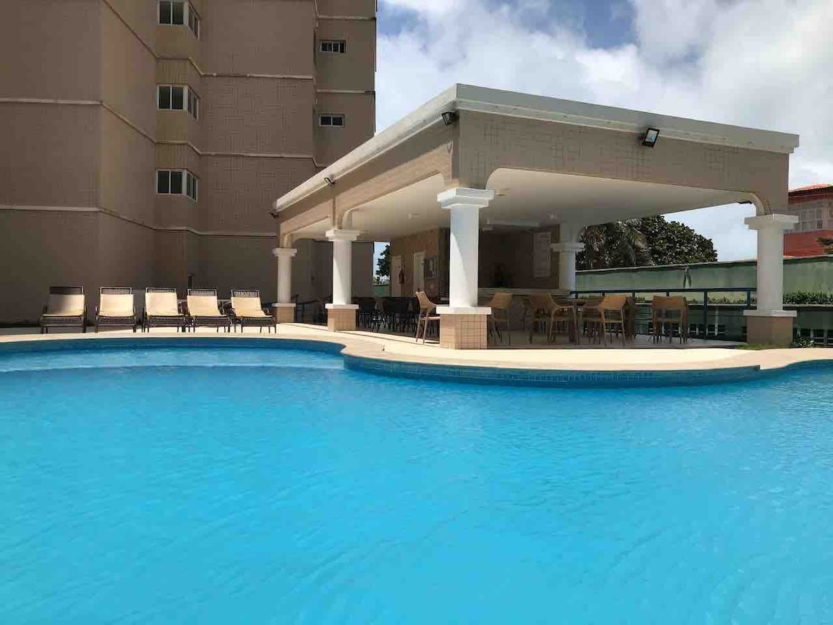 A spacious swimming pool is surrounded by a clean, tiled deck. Lounge chairs are arranged neatly along the edge, with shaded seating areas under a large pavilion. The building's structure is visible in the background, complemented by a clear sky.