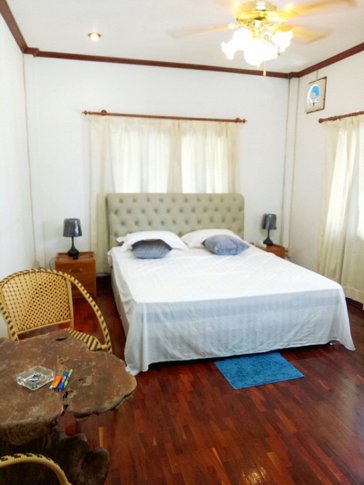 A simple bedroom features a spacious bed with two pillows and a light coverlet. Beside the bed, two lamps are placed on wooden nightstands. A small table made from natural wood adds a touch of rustic charm. Large windows with light curtains allow natural light to fill the room.