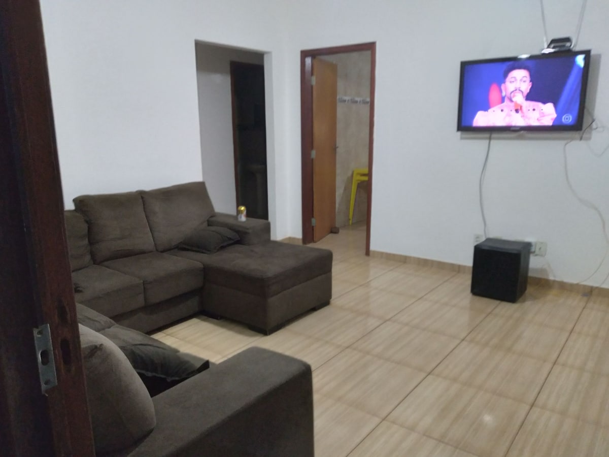 A comfortable living area displays a large brown sectional sofa arranged around a low coffee table. A television is mounted on the wall, providing entertainment. The tiled floor and neutral wall color create a simple and clean atmosphere, complemented by a door leading to another room.