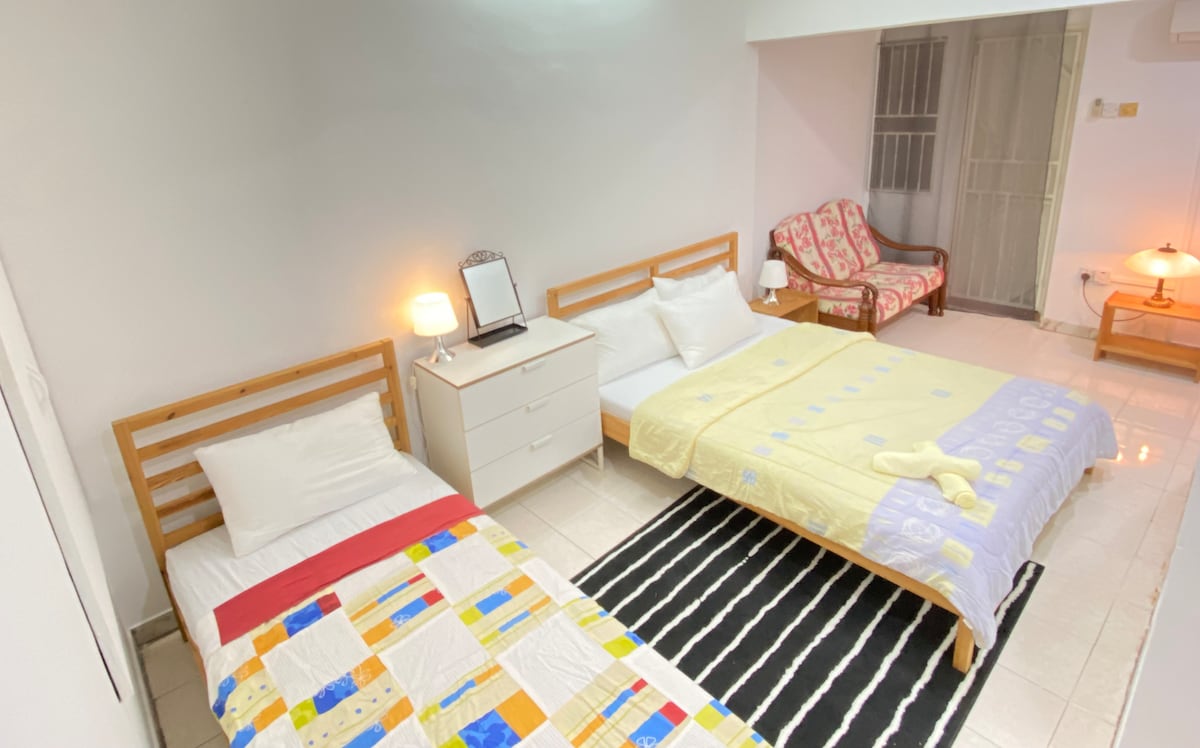 A spacious room is furnished with two comfortable beds, one with a colorful quilt and the other in soft yellow. A wooden nightstand and dresser are positioned nearby, while a cozy seating area is created with armchairs and soft lighting in the corner.