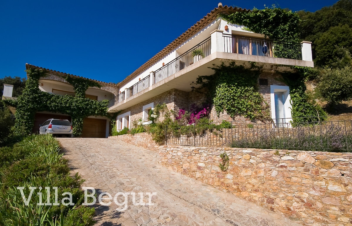 The villa features a two-story design with a natural stone façade, surrounded by lush greenery and vibrant flowers. A driveway leads up to the entrance, where a parked car is visible. Balconies overlook the serene landscape, enhancing the villa's picturesque setting.