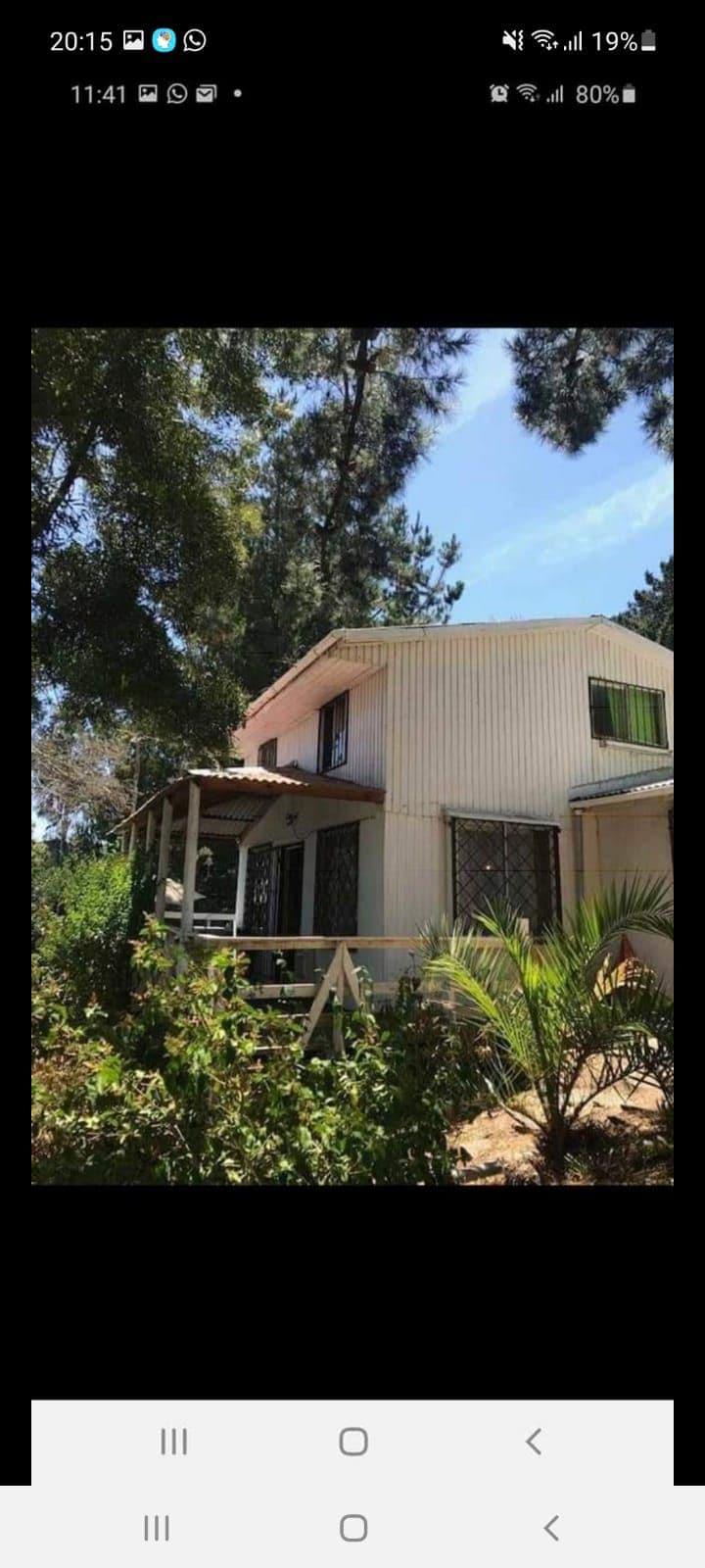 The exterior of a two-story cabin is surrounded by lush greenery, showcasing a blend of natural and architectural elements. A covered porch with steps leads to a welcoming entrance. Large windows offer views of the surrounding trees, contributing to a serene atmosphere.