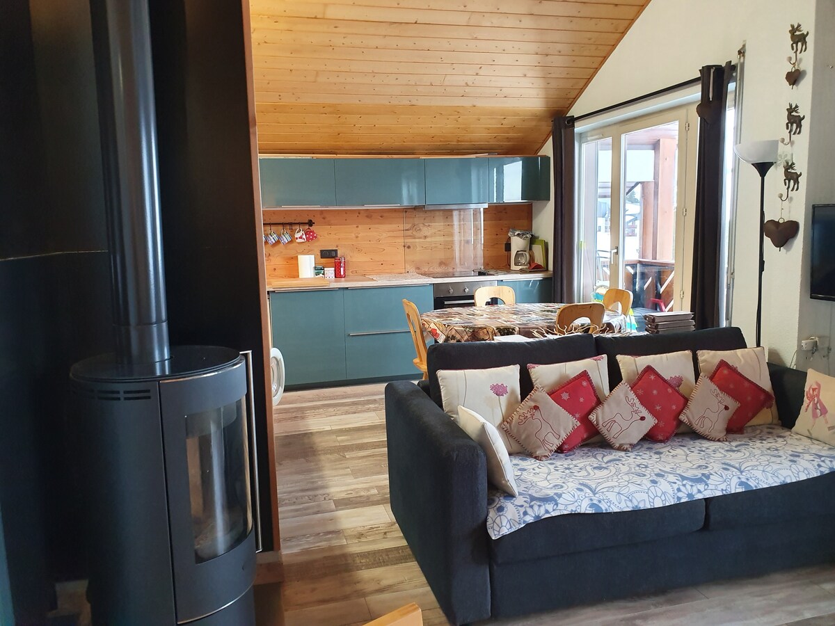 An open living space features a dark sofa adorned with decorative pillows, positioned near a contemporary wood-burning stove. A sleek kitchen with blue cabinetry and wood accents is visible in the background, with ample natural light streaming in from large windows that overlook a balcony.