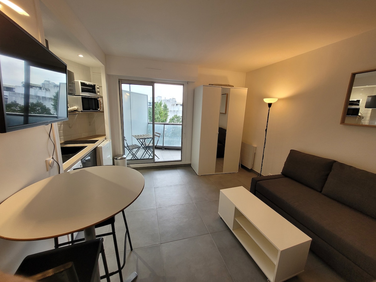 An open living area includes a sleek gray sofa and a white coffee table, complemented by a round dining table with black chairs. A glass door leads to a balcony, while modern kitchen appliances are positioned against the wall, and light fixtures provide a gentle illumination.
