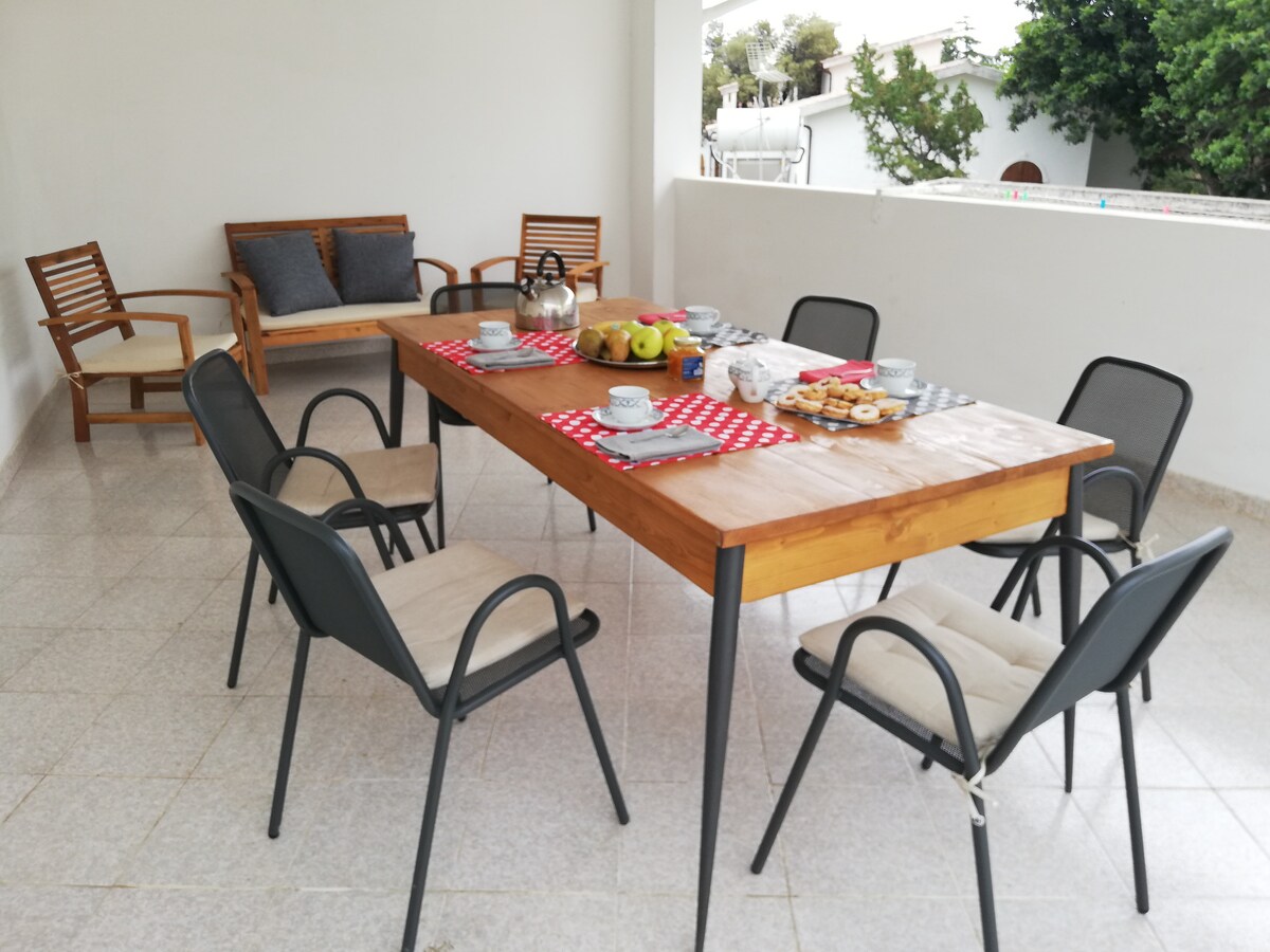 A spacious covered veranda is shown, furnished with a large wooden table surrounded by six chairs. A wooden bench sits against the wall, and a small coffee set is arranged on the table alongside fruit and snacks. Light-colored tiles cover the floor, enhancing the open feel.