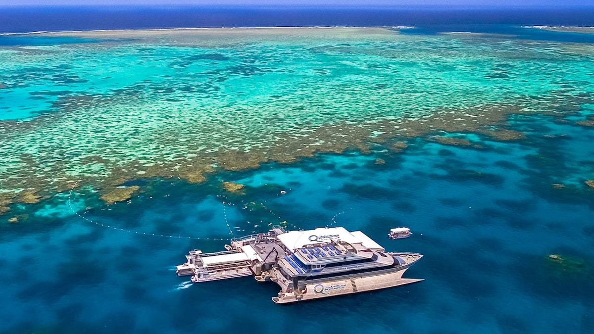 An aerial view captures a large floating facility set in vibrant turquoise waters. Beneath, coral formations stretch out across the ocean, creating a striking contrast with the deep blue. Small boats can be seen near the dock, enhancing the scene of aquatic exploration.
