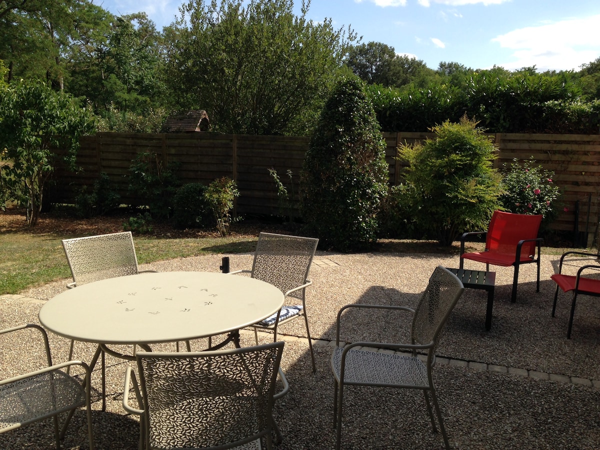 An outdoor terrace features a round table surrounded by four metal chairs, set on a stone surface. Two red chairs are positioned nearby. Lush greenery and vibrant shrubs line the perimeter, providing a natural privacy screen, while a clear blue sky is visible above.