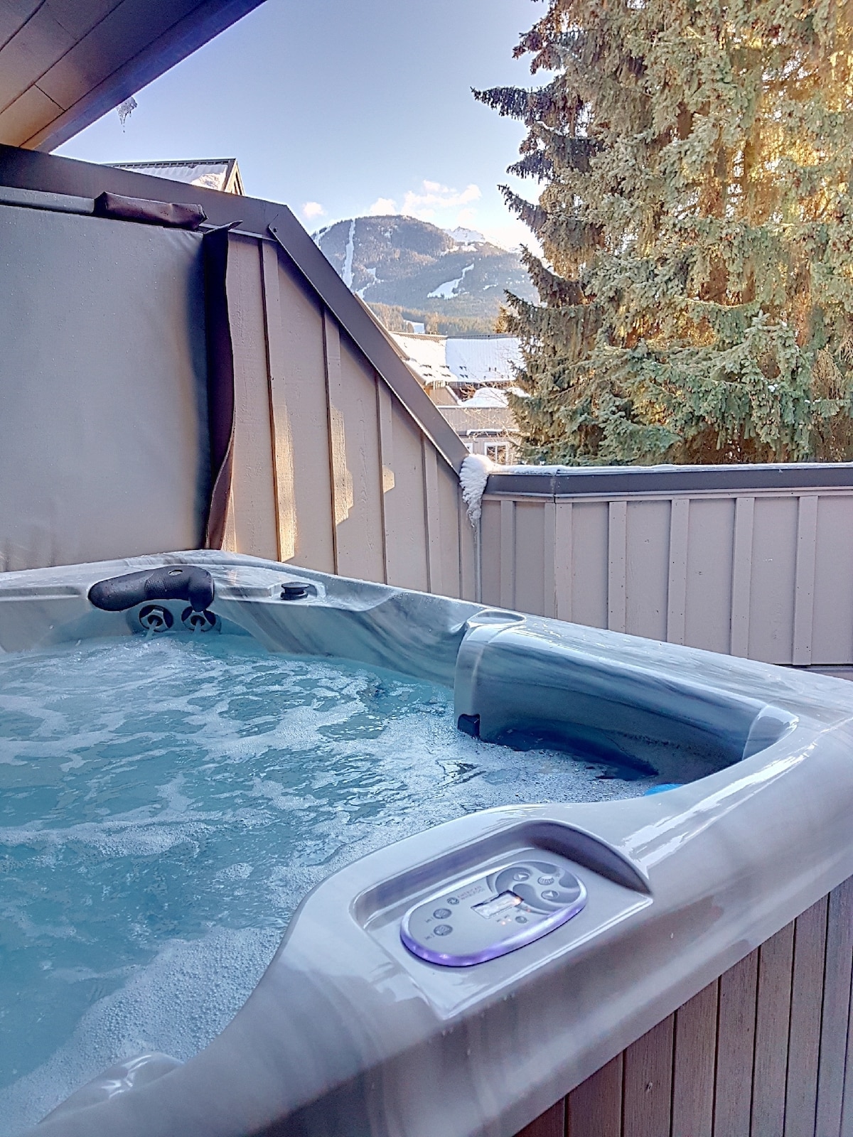 A private hot tub is situated on a deck, with mountain views visible in the background. Sunlight filters through tall trees, creating a serene atmosphere. The hot tub features controls on the side for easy access and relaxation.