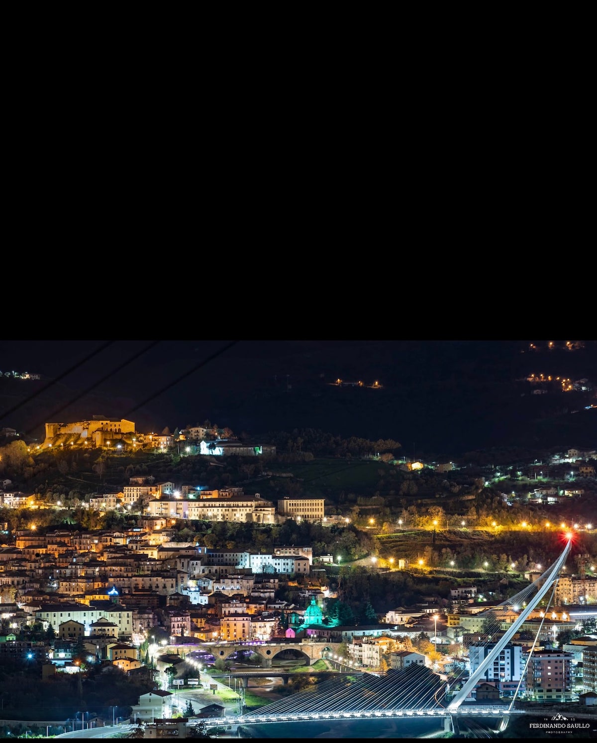 A nighttime view of Cosenza showcases illuminated buildings and winding streets set against a dark sky. The distant hills are outlined by soft lights, creating a serene urban landscape. A prominent bridge is visible in the foreground, leading the eye to the cityscape.