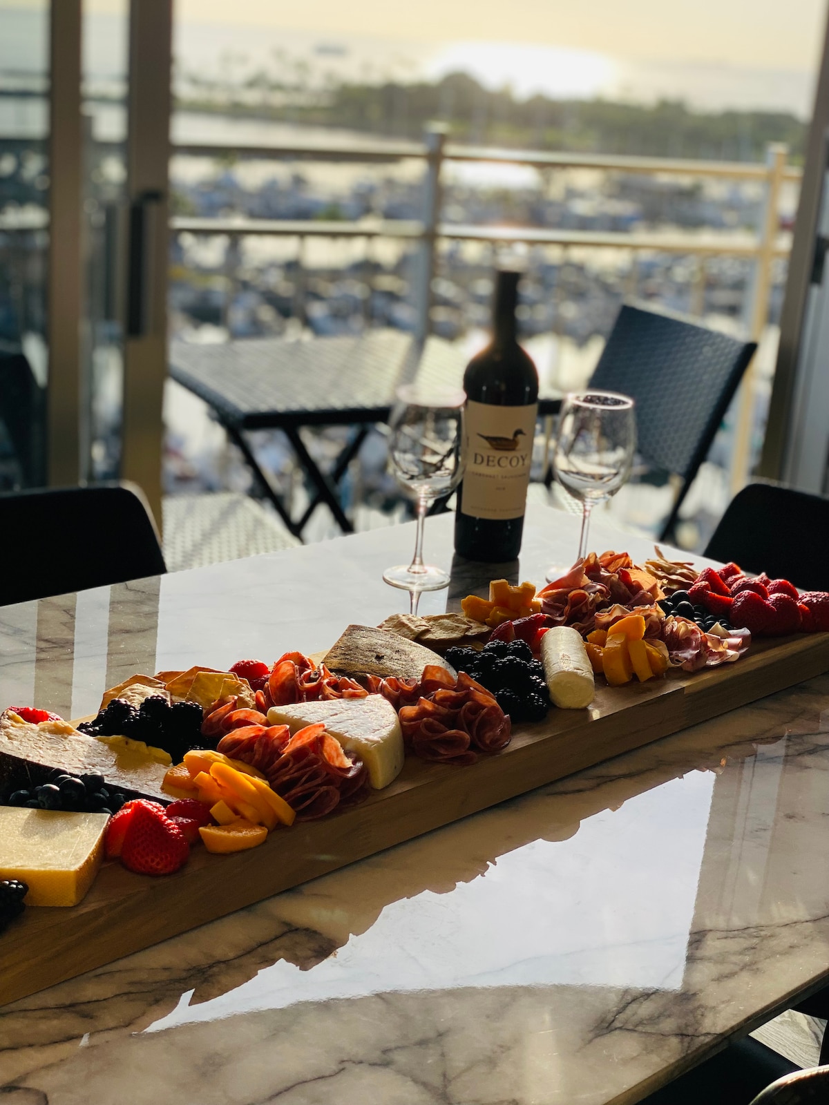A dining table is set with an elegant charcuterie board featuring an assortment of cheeses, cured meats, fruits, and nuts. A bottle of wine and two glasses stand nearby. The background showcases a scenic view of the marina and sunset through large glass doors.