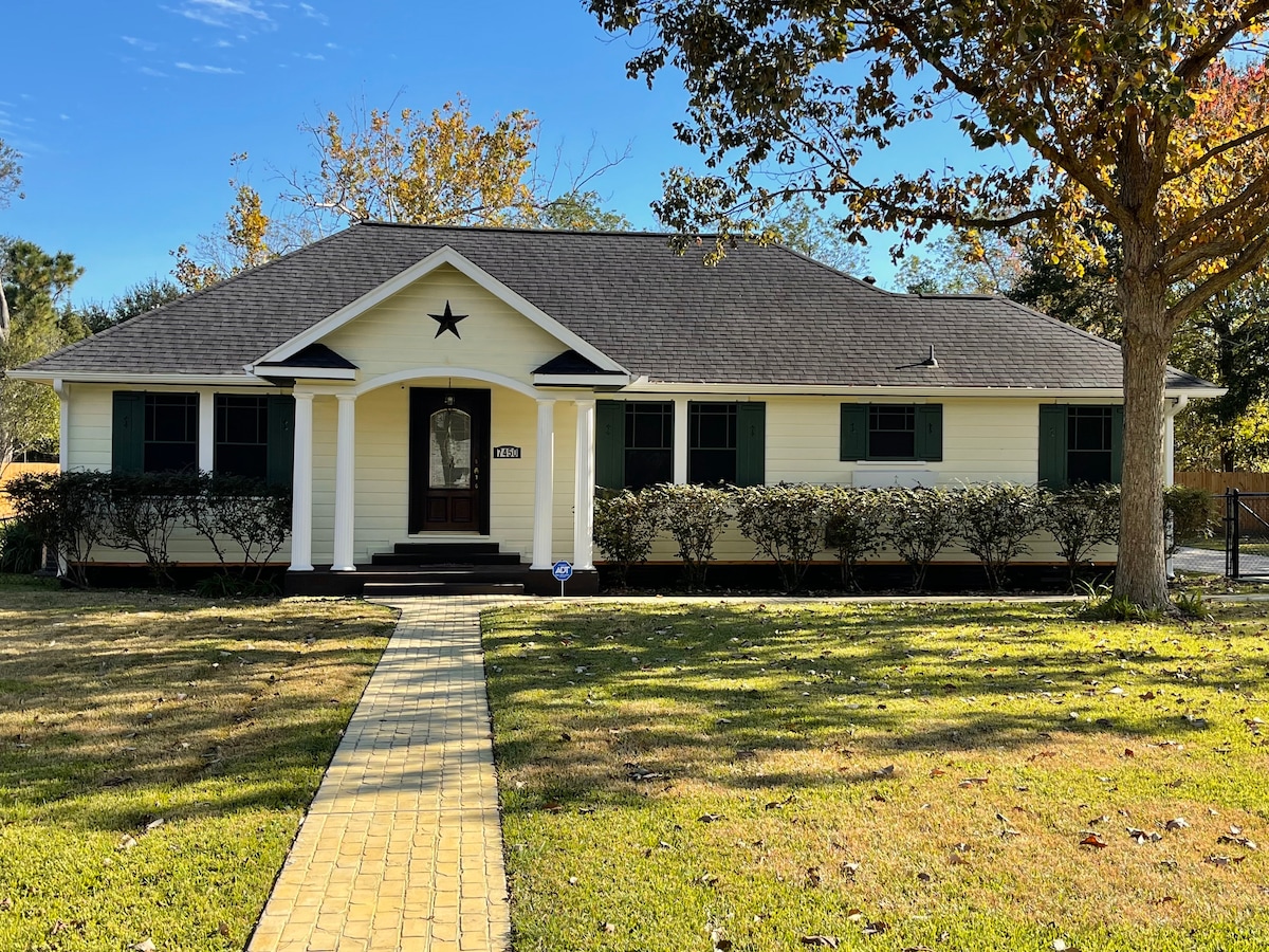 A charming single-story home features a light-colored facade with dark green shutters and a star accent above the front door. A winding yellow brick pathway leads to the entrance, surrounded by neatly trimmed hedges and a spacious lawn adorned with fallen leaves.