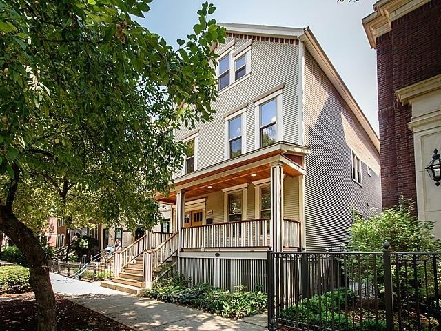 A three-story flat displays a freshly renovated façade with light-colored siding and a welcoming porch. Surrounding trees provide greenery, and a pathway leads to the entrance. The building sits in a well-kept neighborhood, offering a charming atmosphere.