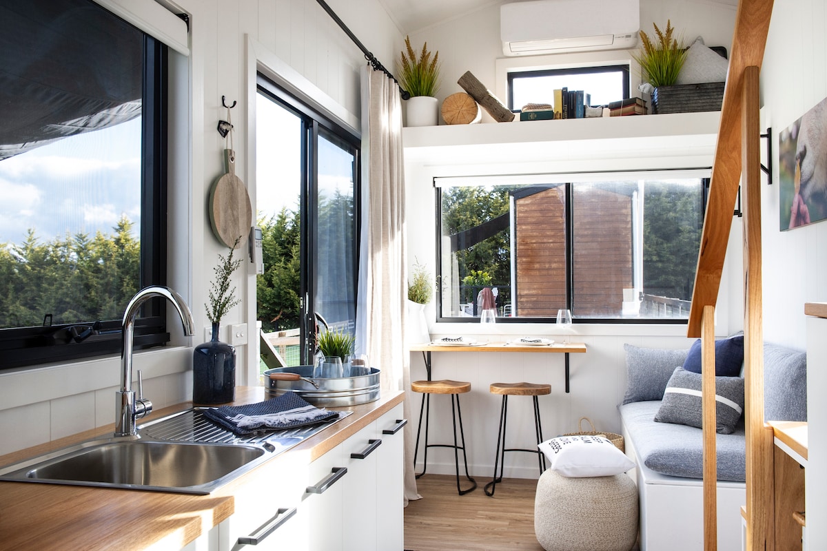 The interior features a well-equipped kitchenette with a sleek sink and wooden countertops. Natural light fills the space through large windows, illuminating a cozy seating area with a comfortable cushion. A small dining space with two stools and a decorative shelf enhances the minimalist design.