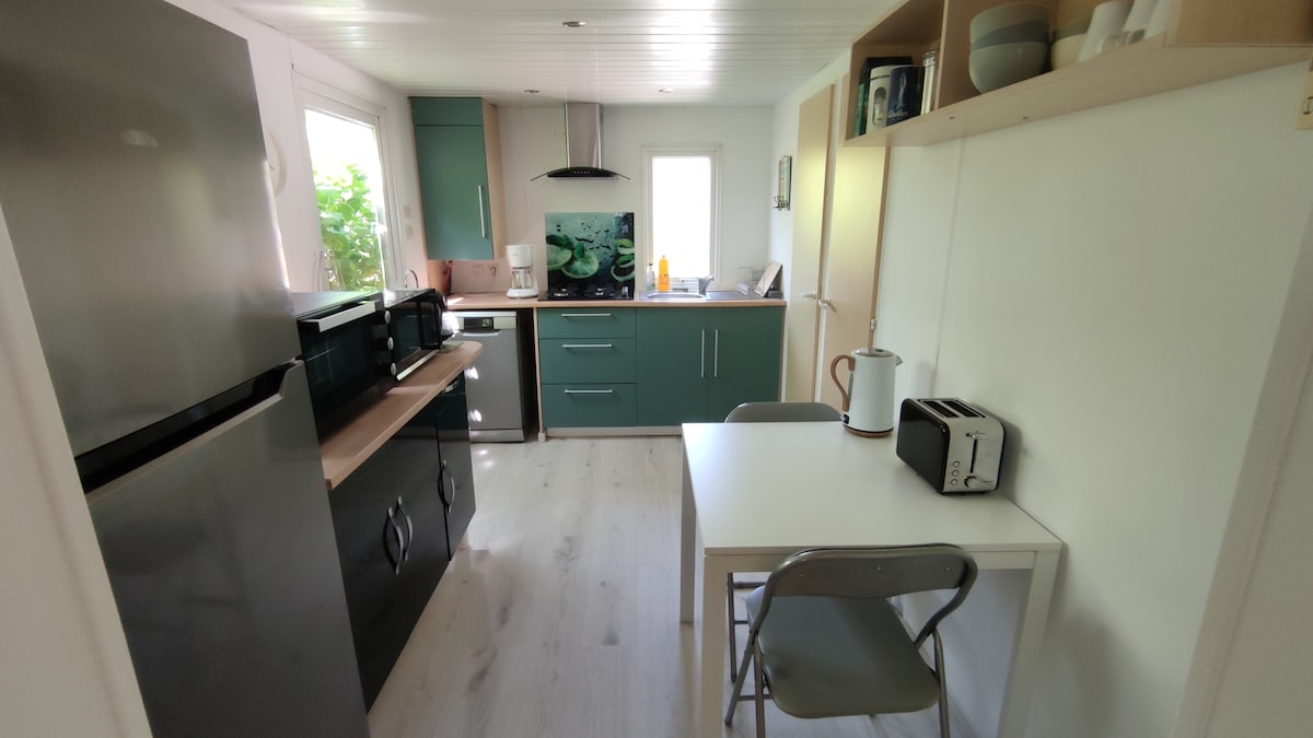 A modern kitchen features sleek cabinets in a muted green tone, complementing the light-colored countertops. Stainless steel appliances include a refrigerator and oven. A small dining table is positioned beside a window, with chairs arranged for seating. Natural light illuminates the space from the adjacent window.