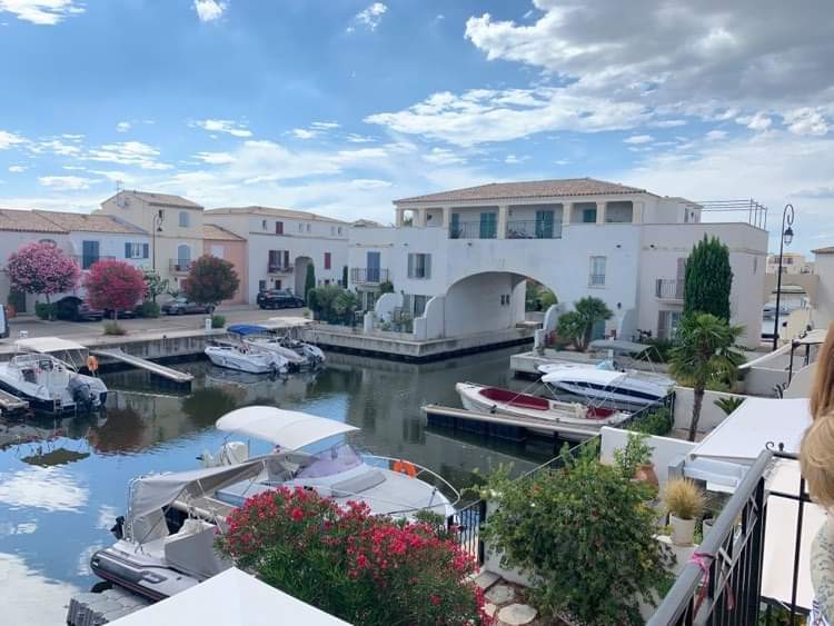 A peaceful waterfront view is presented, featuring several boats moored along the canal. Colorful houses with gardens are visible in the background, complemented by blooming flowers. The sky, adorned with scattered clouds, reflects a serene ambiance over the scene.