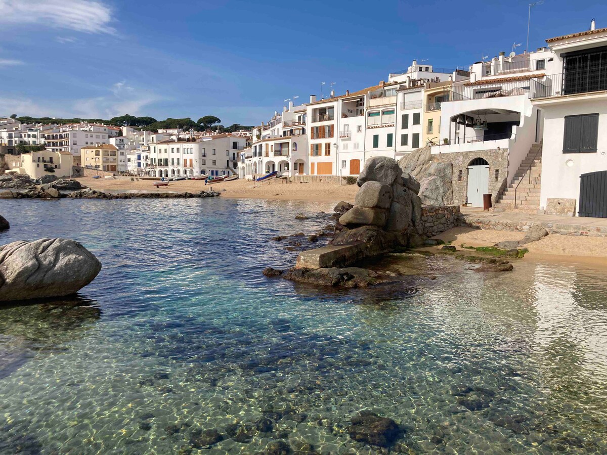 A view of a calm beach area featuring clear turquoise waters surrounded by rocky formations. In the background, colorful buildings line the shoreline, reflecting the coastal atmosphere. The sandy beach curves gently, inviting leisurely walks along the water's edge.