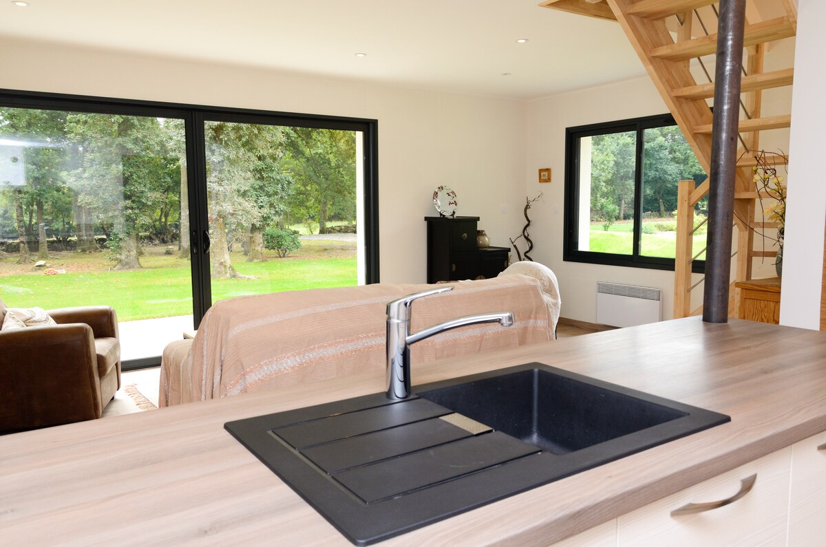 A modern kitchen area features a dark sink positioned in front of a wooden countertop. Large glass doors open to a scenic garden, allowing natural light to fill the space. Comfortable seating and a staircase are visible in the background, enhancing the open layout.