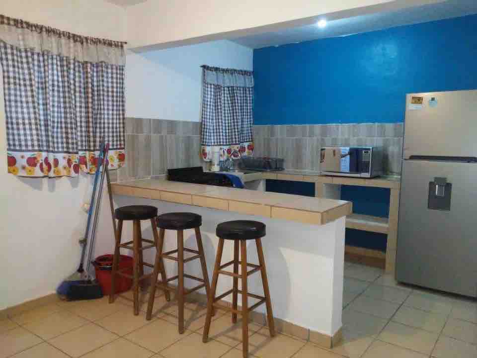 The kitchen area features a bar with three wooden stools and a tiled counter. Blue accent walls complement the checkered curtains over the windows. A silver refrigerator and a microwave are visible, along with basic cleaning supplies on the floor.