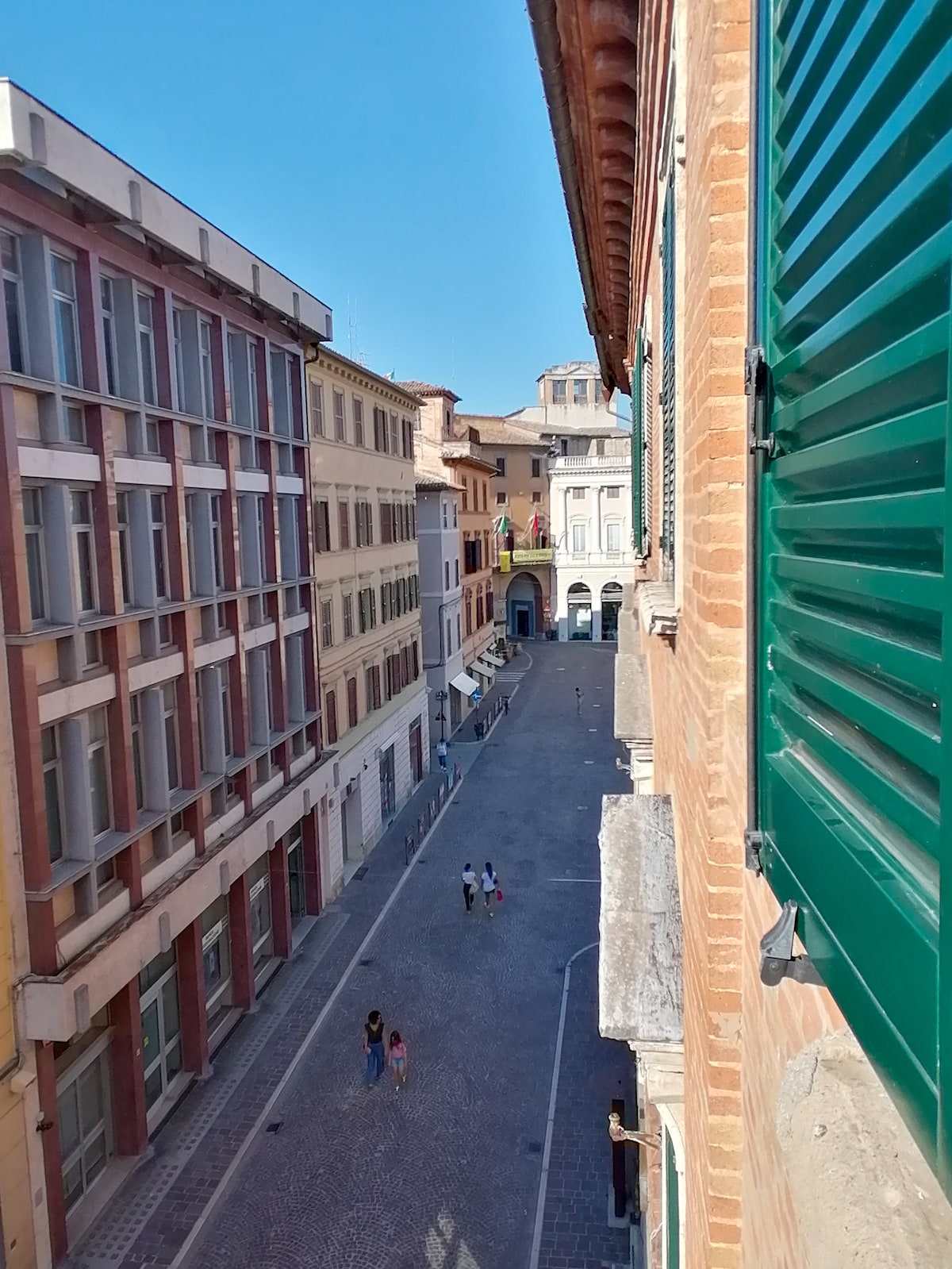 The view from a window is framed by green shutters, showcasing a vibrant street scene below. Cobblestone streets are flanked by buildings of various architectural styles. People can be seen walking along the sidewalk, with a glimpse of shops and historic structures in the background.