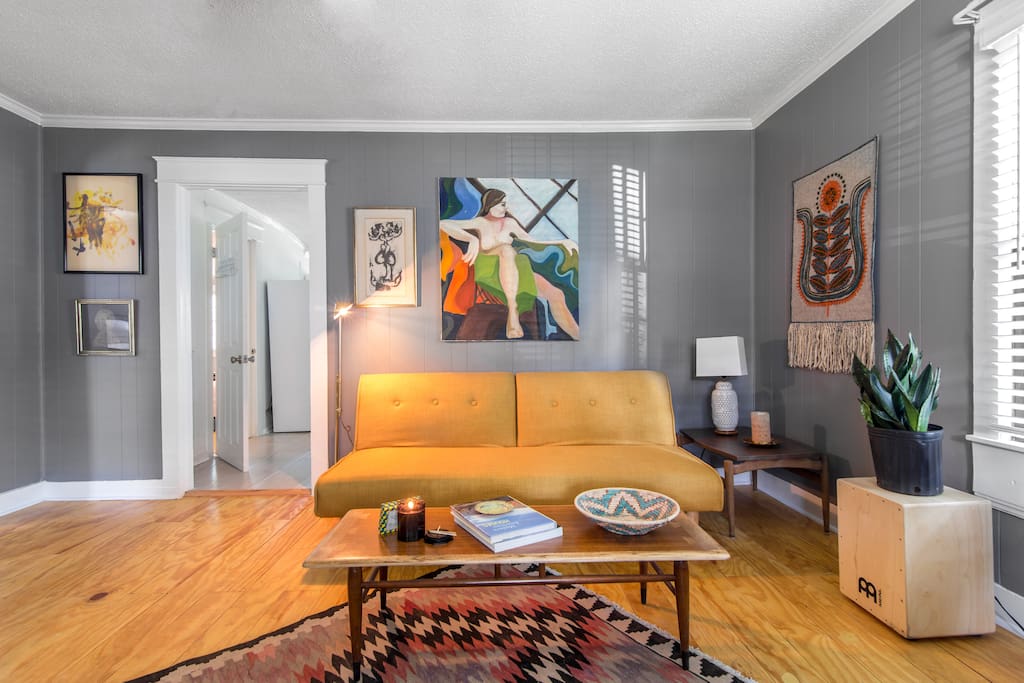 Living room space with Scandinavian light wood floors, collected art and midcentury furnishings. Yellow sofa folds down into a full-sized bed.