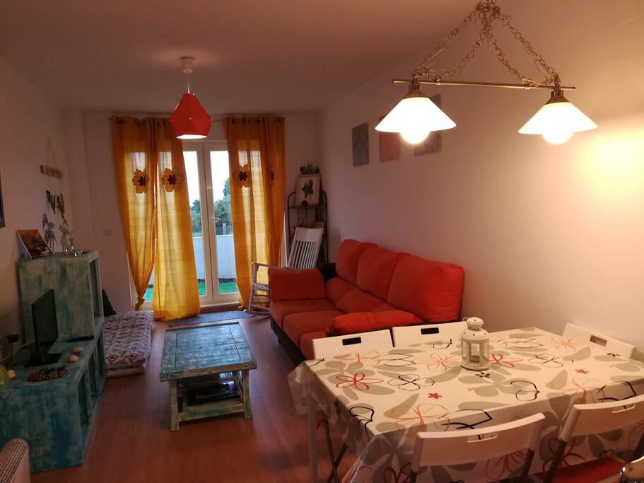 Salón comedor, decoración surfera, con vistas al mar. Sentado en el sofá puedes ver caballos, burros y al fondo el mar. Todo rodeado de prado verde.