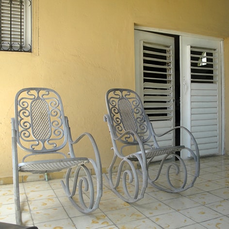 Two metal rocking chairs are positioned on a tiled patio, offering comfortable seating. A light-colored wall serves as the backdrop, while double doors with white shutters provide access to the interior. Natural light bathes the space, enhancing its inviting ambiance.