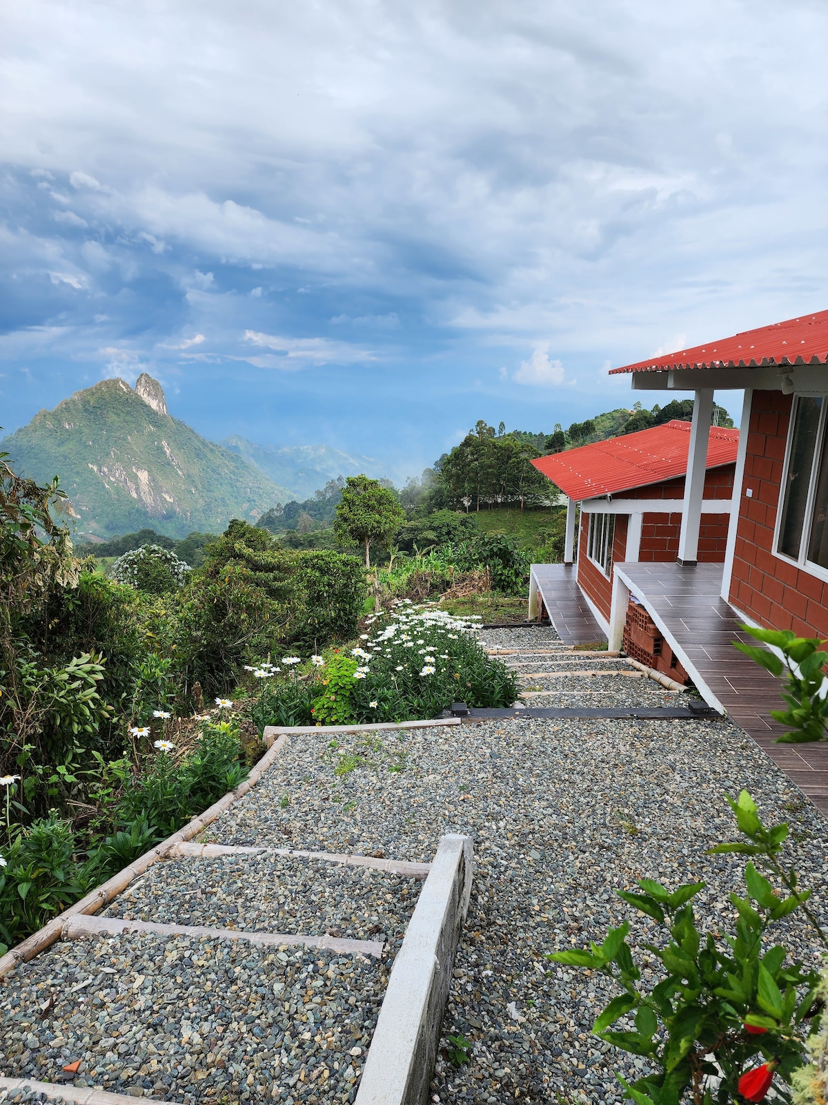 A scenic view captures the hillside retreat, revealing a gravel pathway leading to a cozy red-brick cabin. Lush greenery and blooming white flowers surround the area, and mountains rise majestically in the background under a partly cloudy sky.