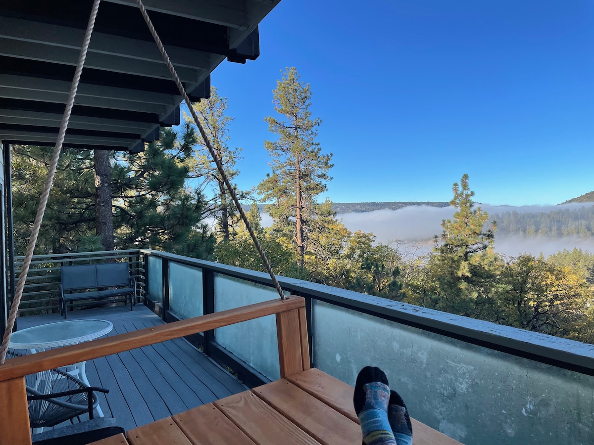 A swing chair is positioned on a deck, offering a view of trees and a misty landscape. The clear sky and distant hills are visible in the background. Two pairs of feet in colorful socks rest comfortably on the deck, indicating a relaxed atmosphere.