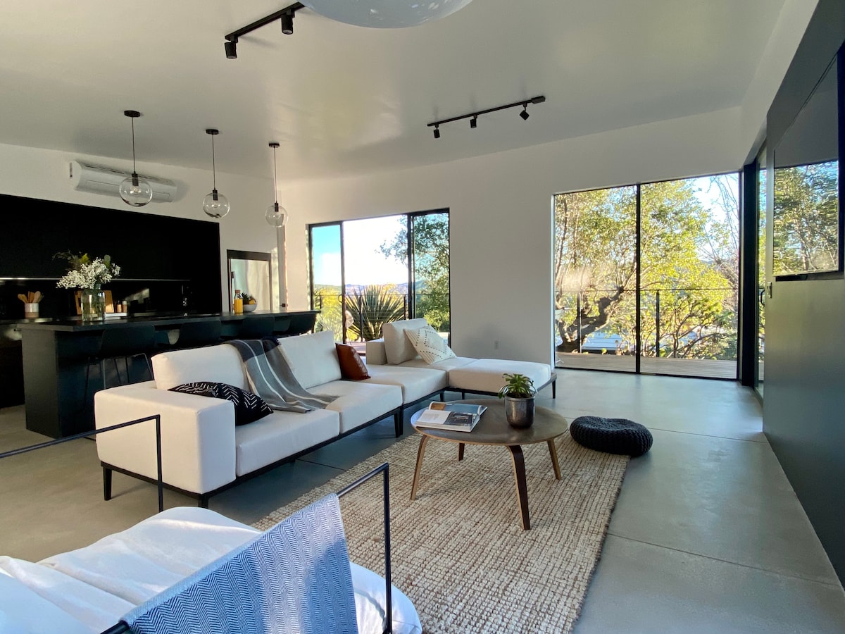 A modern open living space features a comfortable seating arrangement with a sectional sofa and a round coffee table. Large windows allow natural light to fill the area, offering views of the surrounding greenery. A stylish rug adds warmth to the concrete floor.
