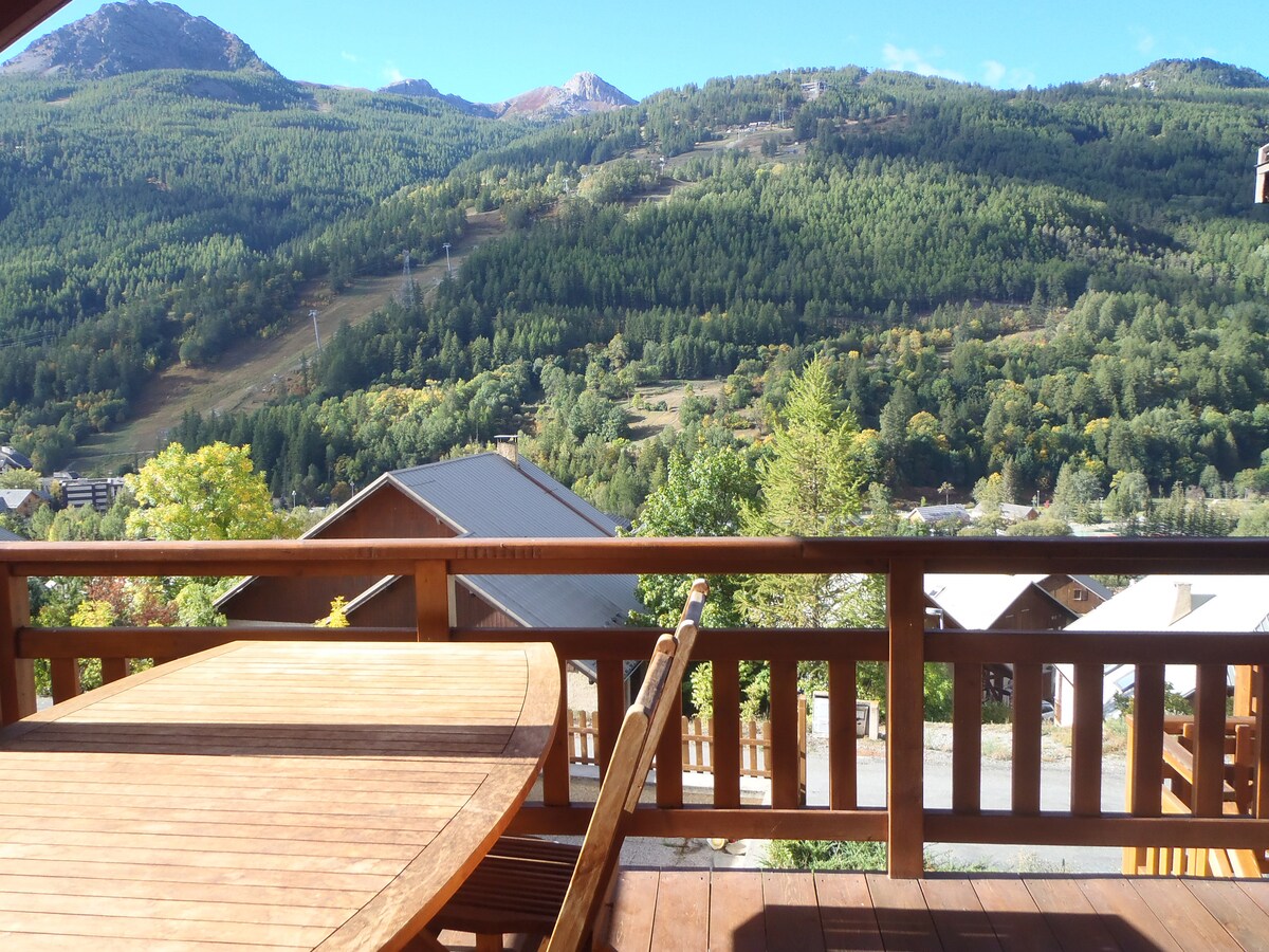 A wooden balcony table and chairs provide a relaxing outdoor space overlooking a lush green mountain landscape. The view showcases a combination of trees and distant hills, with a hint of the ski slopes in the background, embodying the serene alpine setting.