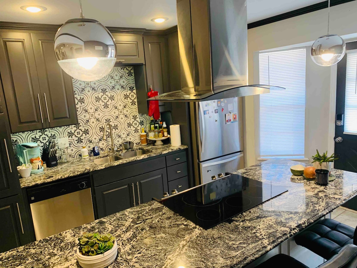 The kitchen showcases modern cabinetry with a dark finish, complemented by a patterned backsplash. A central island features a stovetop and bar seating with two stools. Bright natural light streams through the windows, illuminating the granite countertops and various kitchen essentials along the counter.